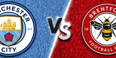 A wide sports matchup graphic featuring Manchester City and Brentford facing off. Manchester City’s crest appears on the left over a textured sky-blue background with faint oversized club branding in the backdrop, while Brentford’s crest is displayed on the right over a textured red background with a faded club logo watermark. A glowing diagonal divider separates the two sides, with large metallic “VS” lettering centered between the clubs. The design uses dramatic lighting, textured patterns, and soft gray borders above and below for a polished promotional match graphic.