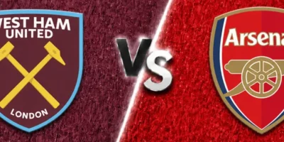 A wide sports matchup graphic featuring West Ham United and Arsenal facing off. West Ham United’s crest appears on the left over a textured claret background with faint oversized club branding in the backdrop, while Arsenal’s crest is displayed on the right over a textured bright red background with a faded club logo watermark. A glowing diagonal divider separates the two sides, with large metallic “VS” lettering centered between the clubs. The design uses dramatic lighting, textured patterns, and soft gray borders above and below for a polished promotional match graphic.